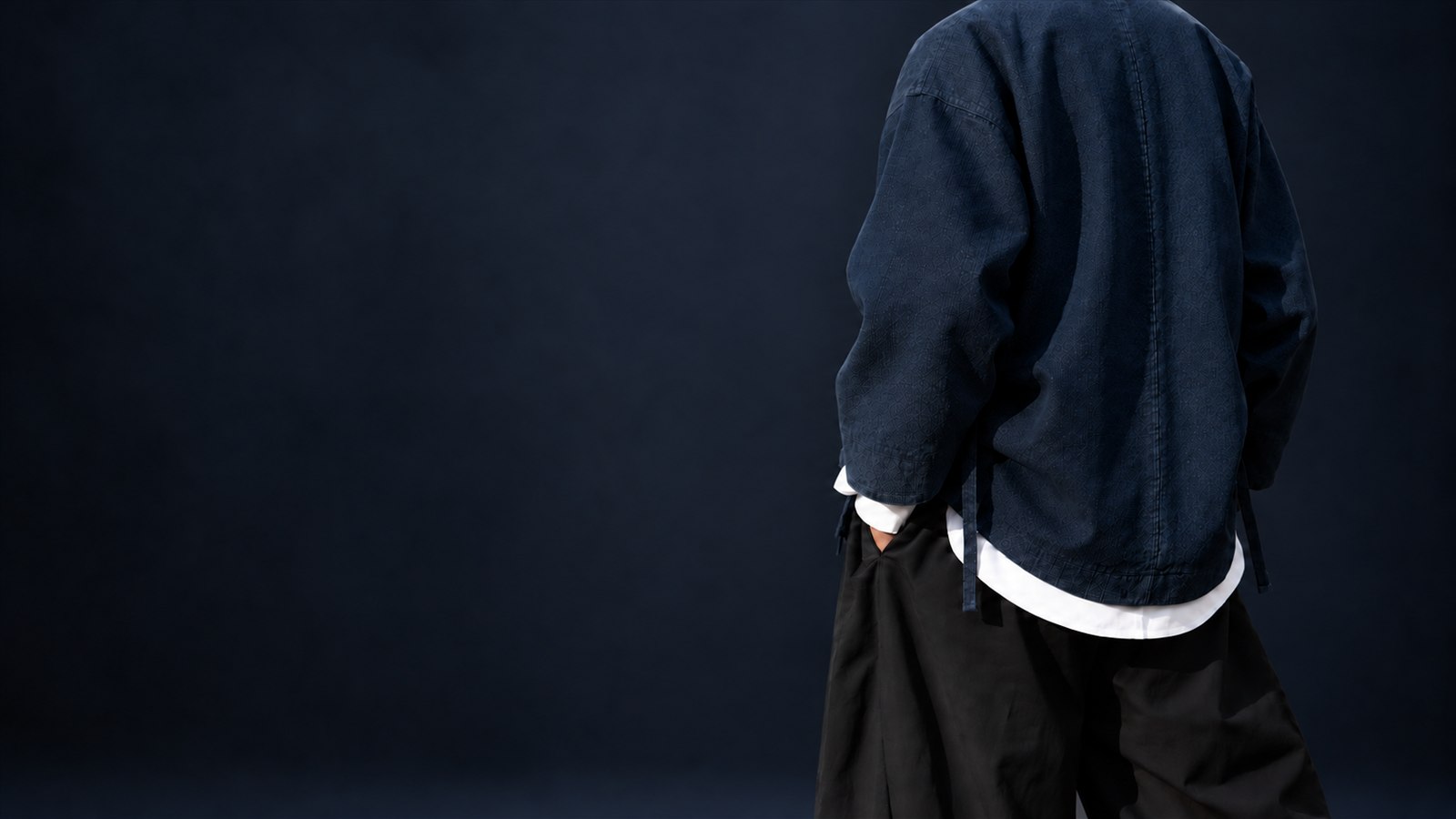 Cropped rear view of a textured navy jacket, white layers and loose black trousers in a dark studio